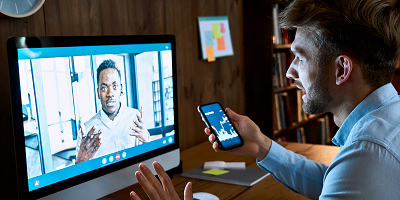 Client video conferencing with financial advisor on computer while holding phone that shows financial graphs
