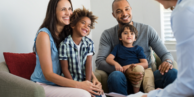 Smiling mixed race family sitting in chairs with kids on parents laps across from life insurance agent
