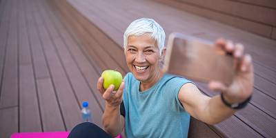Smiling senior woman in athletic clothes holding an apple takes a selfie with a smartphone 
