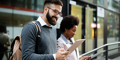 Smiling business man on tablet standing outside building next to female coworker using a smartphone
