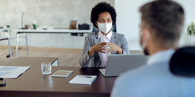 Female financial professional wearing mask sitting at desk with laptop across from male client