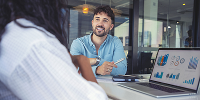 Male financial professional sitting at table with tablet and notebook talking to female client using laptop