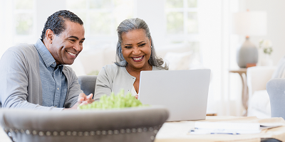 Smiling senior couple sitting at dining room table and video chatting on laptop