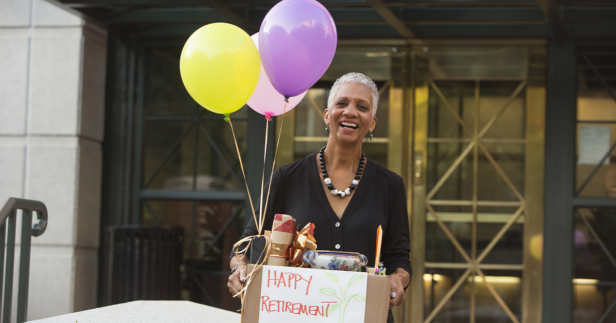 Smiling older woman holding balloons and a box of retirement gifts outside office building
