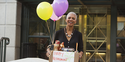 Smiling older woman holding balloons and a box of retirement gifts outside office building