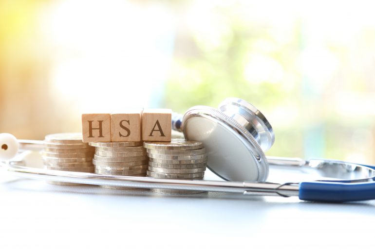 Scrabble tiles spelling out “HSA” displayed on stack of coins and with stethoscope. 