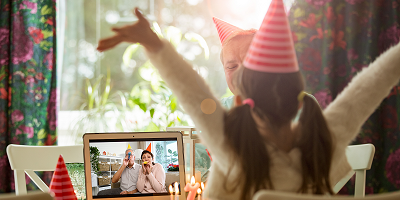 Young girl wearing party hat throws hands in the air over birthday cake with grandparents on laptop screen 